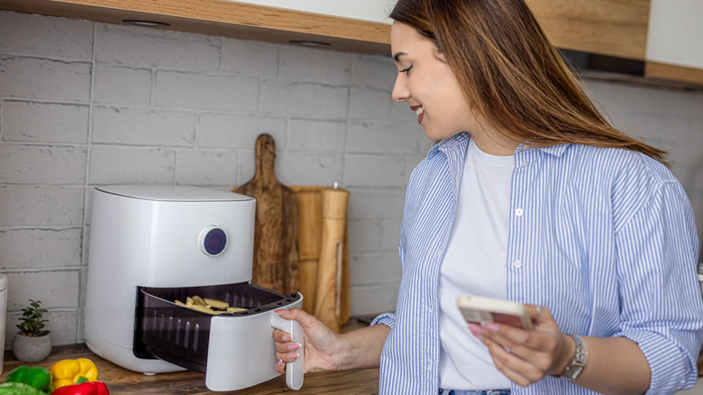 Young woman adjusting settings on a compact air fryer while holding a smartphone in a stylish home kitchen