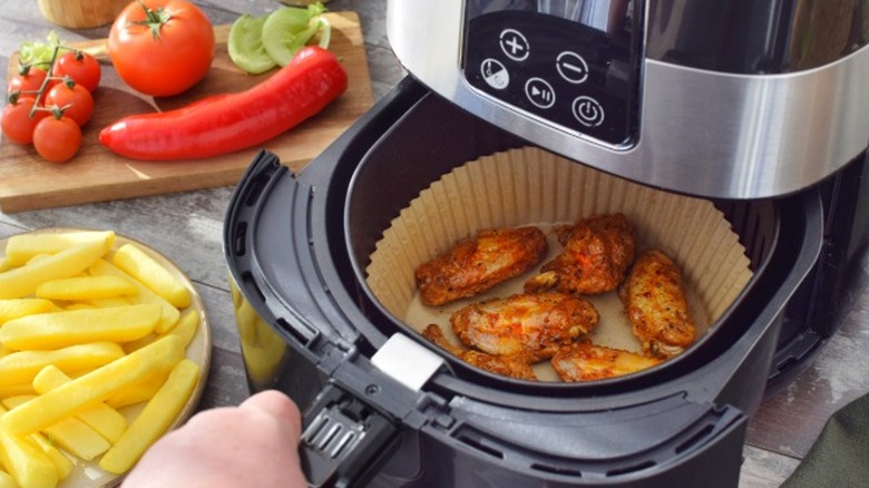 Chicken wings being prepared in an air fryer with a liner on a countertop next to some cut potato fries and tomatoes.