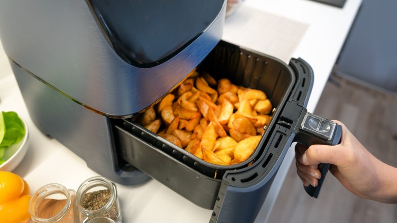 An air fryer on a kitchen counter with a hand pulling the slide-out drawer open to reveal freshly made potato wedges.