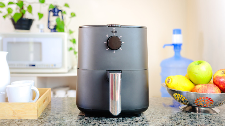 An air fryer on top of a counter top next to a bowl of fruit and a tray of drinks