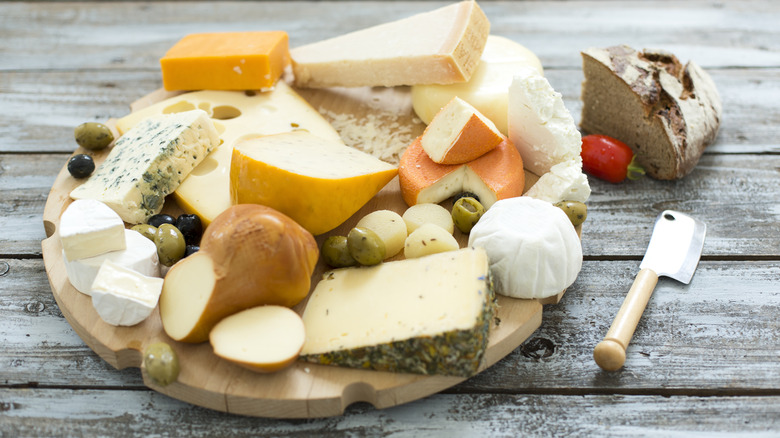 A round cutting board with various cheeses and olives on a wooden tabletop.