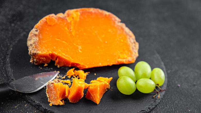 A piece of mimolette on a black plate with a cheese knife and green grapes.