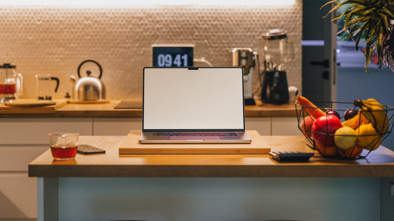 An open laptop is seen on a kitchen counter beside items like a fruit bowl and a measuring cup