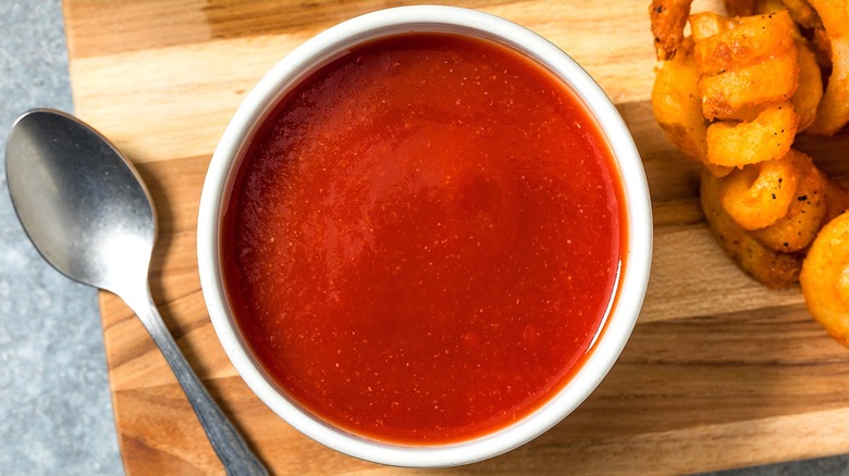 A vinegar-based barbecue sauce is in a white bowl next to curly fries and a metal spoon