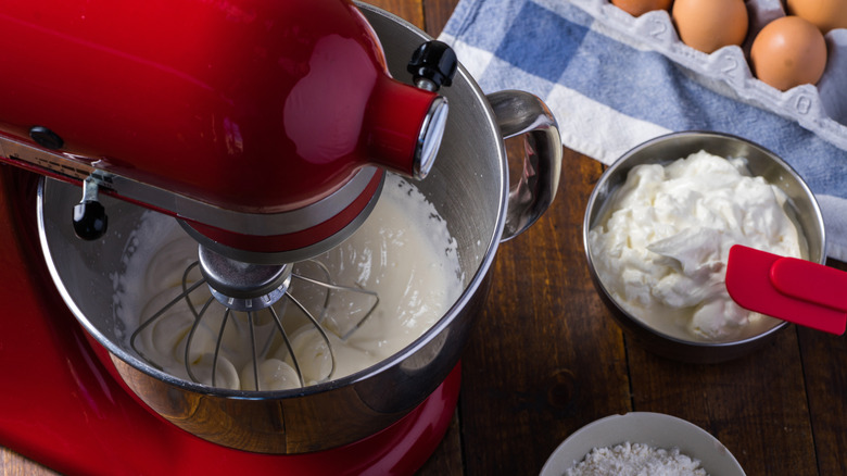 Red stand mixer whipping cream near eggs and several bowls of ingredients.