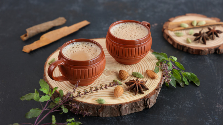 Two cups of chai on top of a wooden board over a grey countertop surrounded by chai spices.