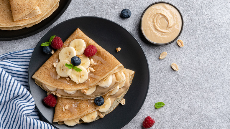 Crepes with peanut butter, banana slices, honey, and fresh berries on black ceramic plate.