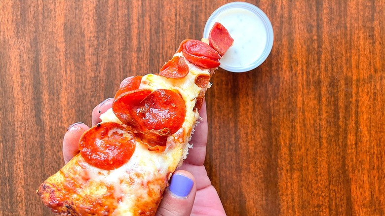 Hand dipping a slice of pepperoni and cheese pizza in a small plastic cup of ranch dressing