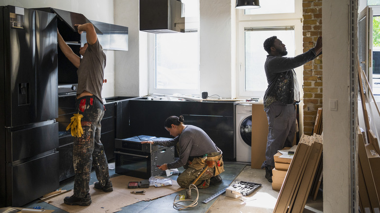 A kitchen being renovated by three workers, two focusing on appliances and one tending to the wall