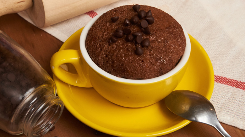 Mug brownie in a yellow cup on a yellow saucer topped with chocolate chips, with a kitchen towel, rolling pin, and mason jar of chocolate chips in the background.