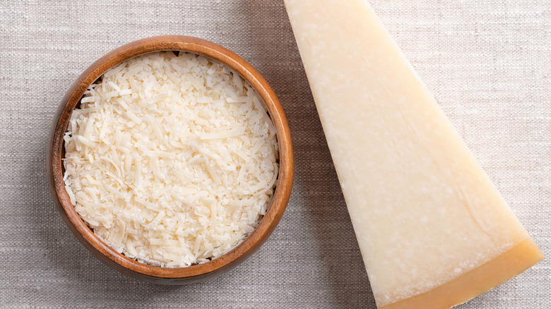 A small bowl of grated Parmesan cheese next to a wedge of Parmesan cheese.