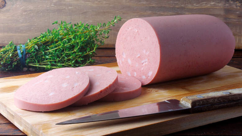 A large "tube"of bologna with three thick slices cut off of it on a wooden cutting board with a knife, a bundle of thyme in the background.
