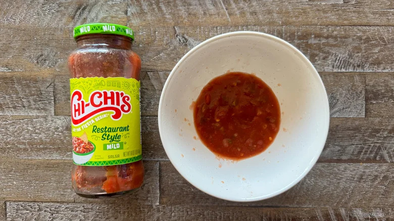 A jar of Chi-Chi's Salsa on a table with some poured out into a bowl next to it.