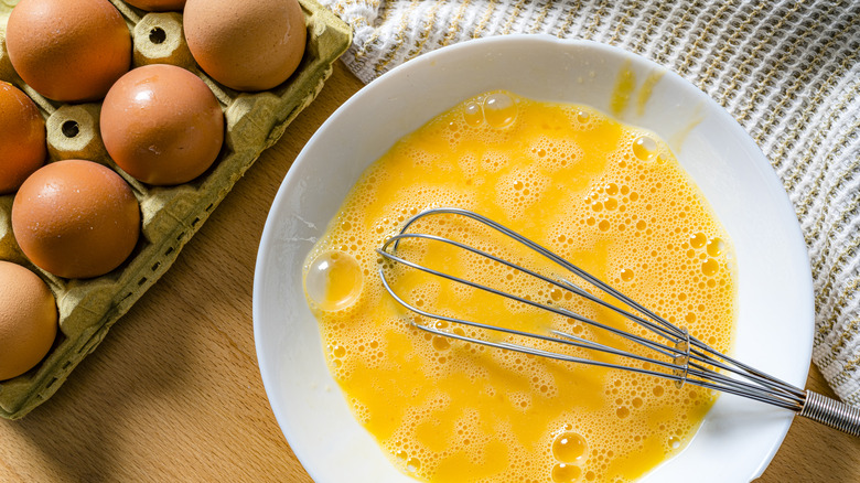 Eggs in shells next to cracked eggs in a white bowl with whisk