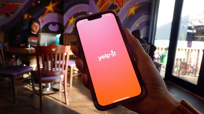 A person's hand is seen holding a phone displaying the Yelp app in a restaurant setting.