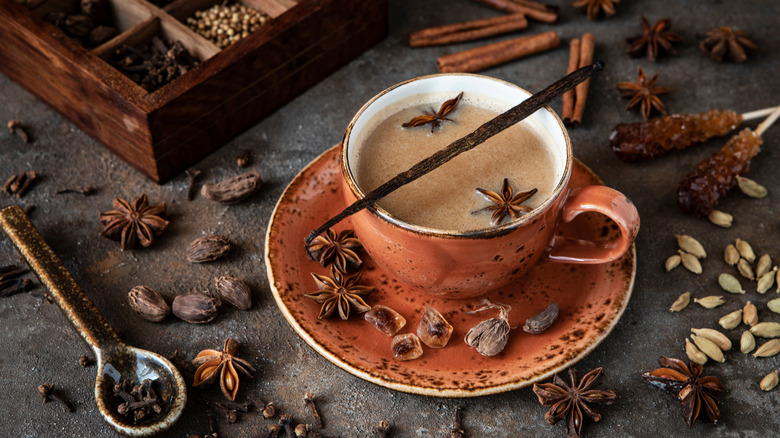 A cup of masala chai with ingredients above. Cinnamon, cardamom, anise, sugar over dark texture background