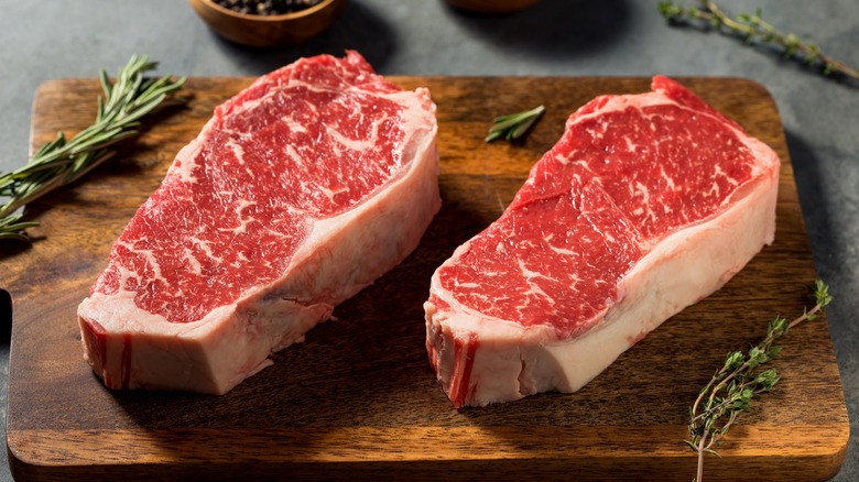 two raw new york strip steaks with thick fat caps on a wooden board