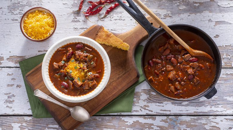 A big pan of chili sits next to a large bowl of chili and a slice of cornbread and cheese