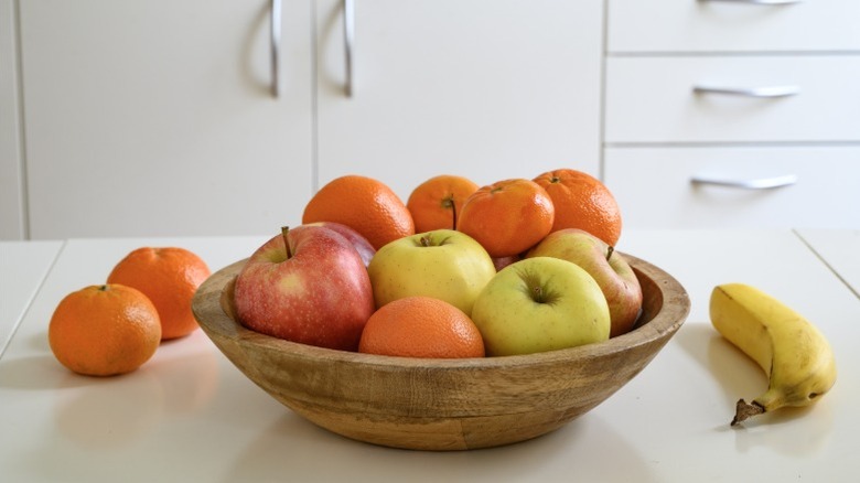A bowl of fresh fruit on a white kitchen counter with a banana and two clementines sitting around it.
