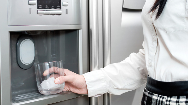 A person holding a glass under a refrigerator door ice machine.