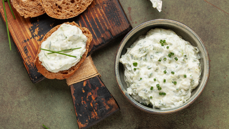 A bowl of Kentucky Benedictine spread, topped with chives next to a cutting board with sliced bread topped with the spread.