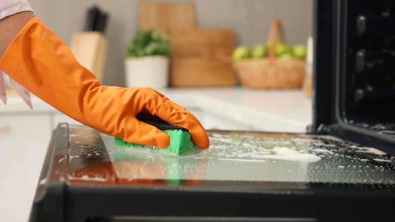 An individual cleaning an oven door with a sponge.