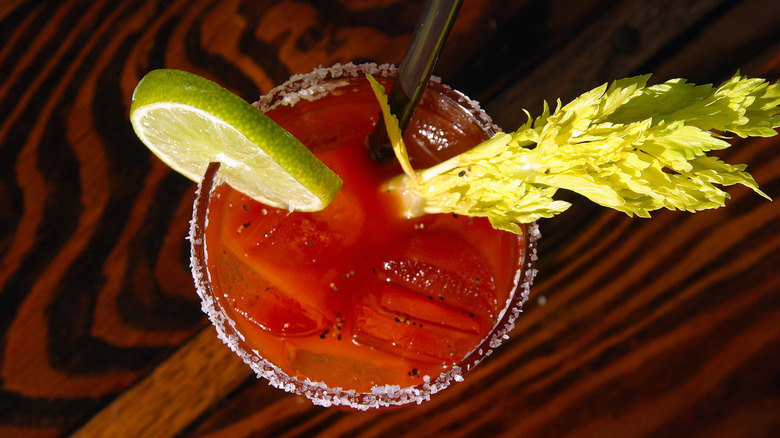 Bloody Mary cocktail garnished with celery, okra, onion, olive and salt rim on a rustic wooden table. A Bloody Mary is a cocktail containing vodka, tomato juice, and other spices.