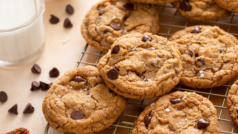 A fresh batch of chewy chocolate chip cookies on a cooling rack with a glass of milk and chocolate chips to the side.