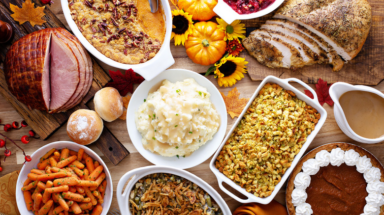 Dinner table laden with Thanksgiving dishes including a plate with turkey, sweet potatoes, green beans, stuffing, and cranberry sauce with utensils and napkins