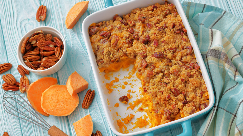 Sweet potato casserole with pecan streusel topping in blue and white dish with servings removed, on blue plaid towel on blue wood table, with sweet potato slices and a cup of shelled pecan halves nearby