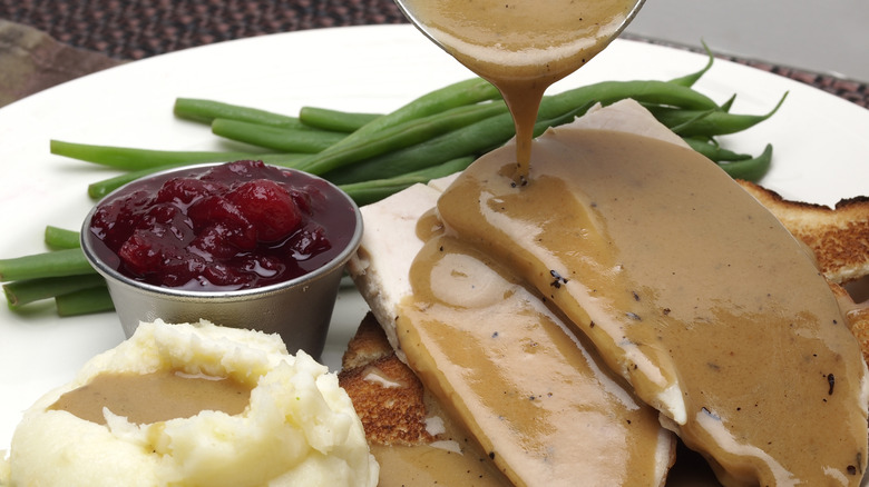 Person pouring gravy over slices of turkey on plate with green beans, mashed potatoes, and cranberry sauce