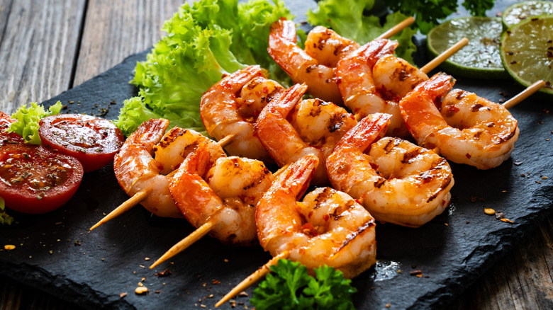 A stone plate with skewers of seasoned shrimp on top of it alongside vegetables