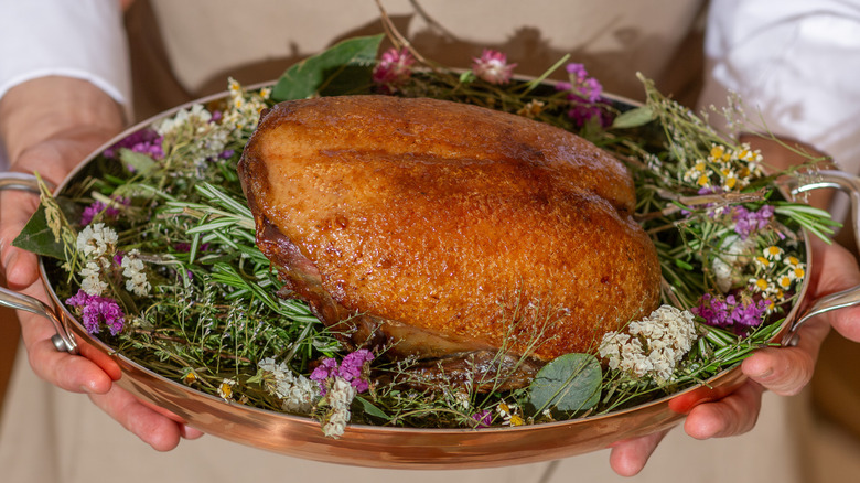 Chef holding a roasted turkey breast in a copper dish surrounded by herbs and flowers