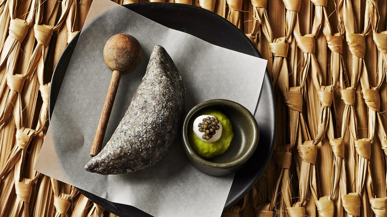 A black corn empanada on a black plate set on a woven mat