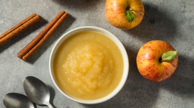 Bowl of appleauce next to apples, cinnamon sticks, and spoons