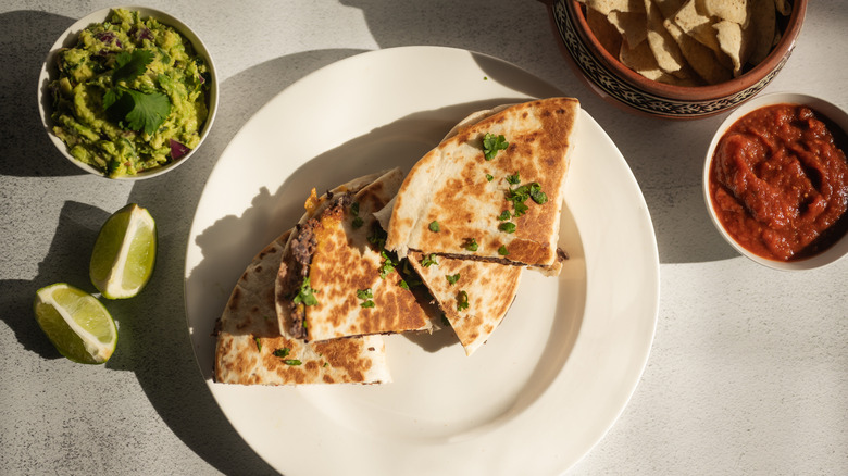 Quesadilla on a white plate with guacamole, salsa, tortilla chips, and lime wedges on the side
